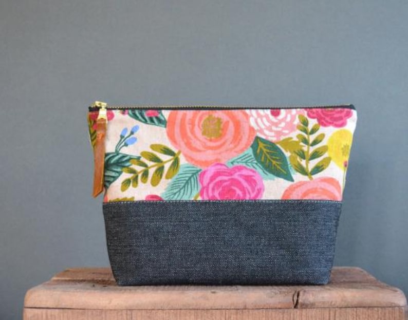 Pu&ograve; includere: Una pochette con zip a stampa floreale con fondo in denim nero. La pochette presenta una variet&agrave; di fiori rosa, rossi, gialli e verdi su uno sfondo bianco.