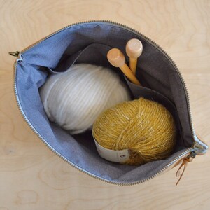 May include: A tan leather zippered pouch with a gray fabric lining. The pouch contains two wooden knitting needles and two balls of yarn, one white and one yellow.