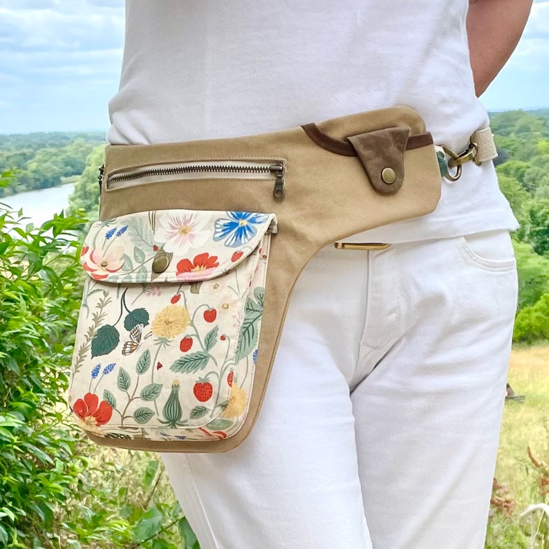 Pu&ograve; includere: Una cintura in tela beige con una tasca a stampa floreale. La tasca ha una chiusura a zip e una patta con chiusura a bottone. La cintura ha un cinturino in pelle marrone e una fibbia in metallo.