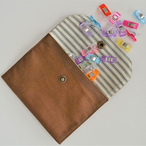 May include: A brown fabric pouch with a striped interior and a snap closure, overflowing with colorful sewing clips. The clips are various colors, including red, pink, purple, and blue. The pouch is open, displaying the contents.