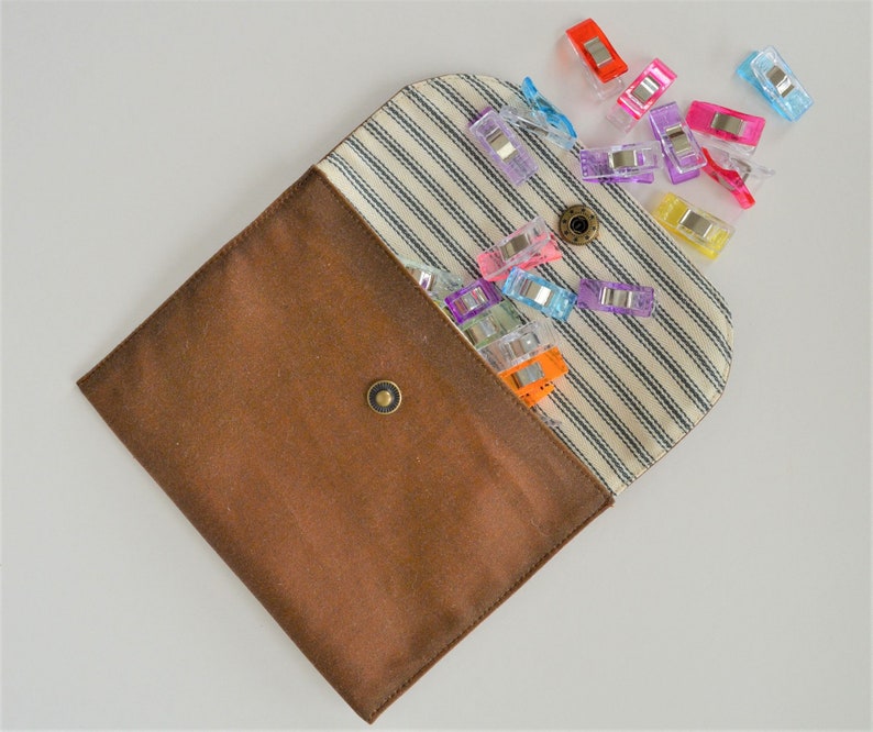 May include: A brown fabric pouch with a striped interior and a snap closure, overflowing with colorful sewing clips. The clips are various colors, including red, pink, purple, and blue. The pouch is open, displaying the contents.