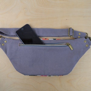 May include: A gray canvas fanny pack with a zippered pocket and a black smartphone inside. The fanny pack has a brown adjustable strap with a metal buckle.