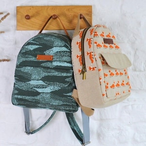 May include: Two small backpacks hanging on a wooden hook. The backpack on the left is teal blue with a white and black pattern. The backpack on the right is beige with a pattern of orange foxes.