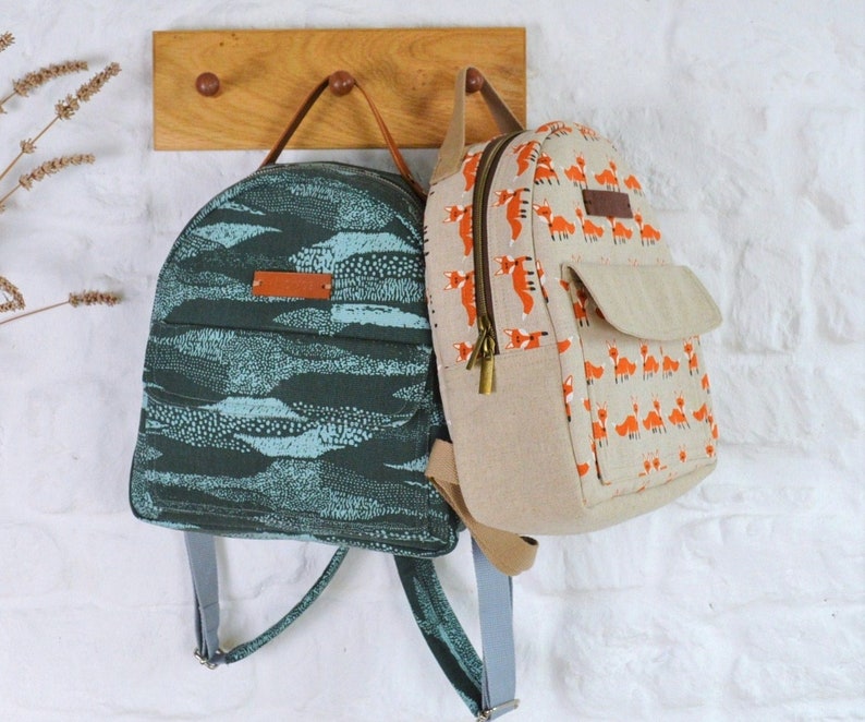 May include: Two small backpacks hanging on a wooden hook. The backpack on the left is teal blue with a white and black pattern. The backpack on the right is beige with a pattern of orange foxes.