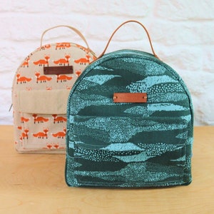 May include: Two small, round backpacks. One is green with a blue and white pattern and the other is beige with a repeating pattern of orange foxes. Both backpacks have a brown leather handle and a brown leather label.
