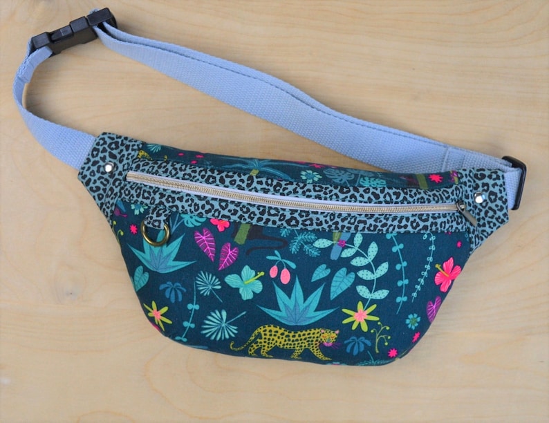 May include: A teal blue fanny pack with a zipper closure and a leopard print trim. The main fabric features a tropical print with green leaves, pink flowers, and a yellow leopard.