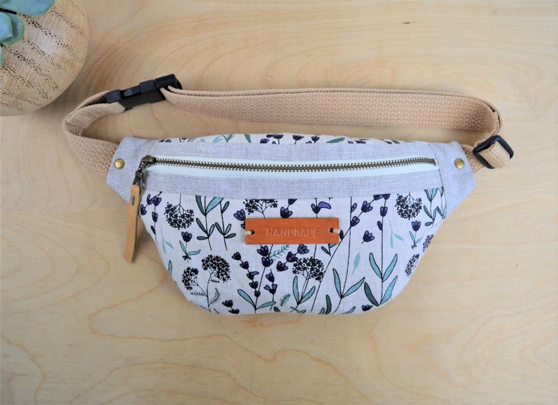 May include: A white fanny pack with a floral print featuring purple flowers and green leaves. The fanny pack has a zipper closure and a brown leather tag that says "HANDMADE".