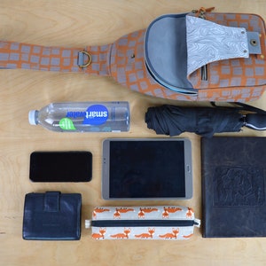 May include: A brown and orange patterned crossbody bag with a gray interior, a black and gray tablet, a black phone, a black leather wallet, a black and white pencil case with a fox print, a clear water bottle with a blue label that says "Smartwater", a black umbrella, and a brown leather journal.