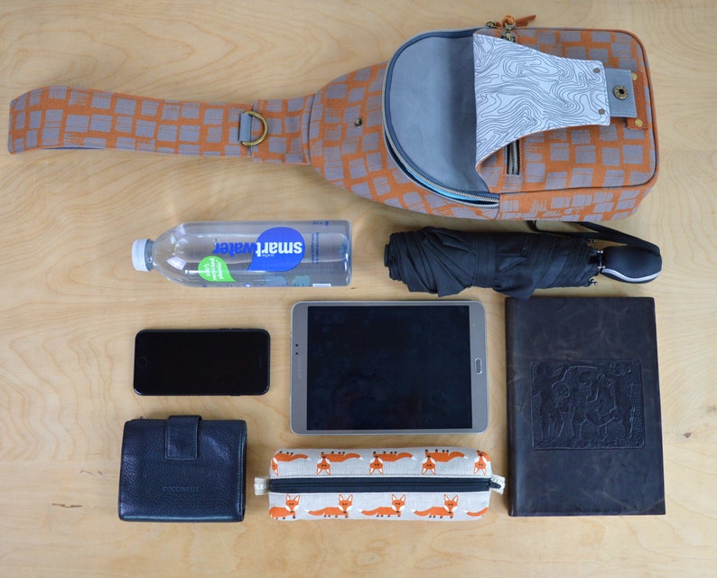 May include: A brown and orange patterned crossbody bag with a gray interior, a black and gray tablet, a black phone, a black leather wallet, a black and white pencil case with a fox print, a clear water bottle with a blue label that says "Smartwater", a black umbrella, and a brown leather journal.