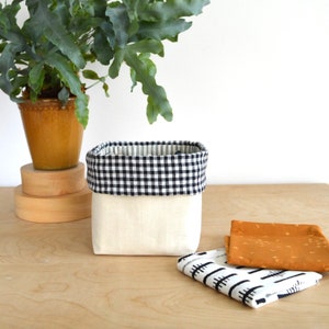 May include: A square, off-white fabric storage basket with a black and white checkered trim. Two small patterned fabric squares are next to the basket. A potted plant sits in the background.