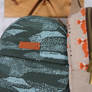 May include: Two canvas backpacks, one with a blue and green abstract pattern and the other with a beige background and a repeating pattern of orange foxes. The backpack with the abstract pattern has a brown leather tag with the word "Canvas" printed on it.