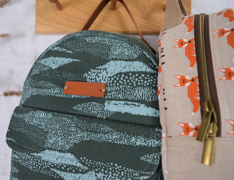 May include: Two canvas backpacks, one with a blue and green abstract pattern and the other with a beige background and a repeating pattern of orange foxes. The backpack with the abstract pattern has a brown leather tag with the word "Canvas" printed on it.