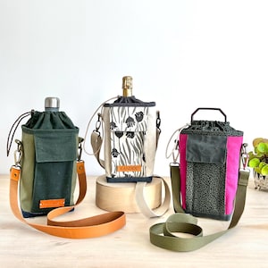 May include: Three cylindrical bottle bags with adjustable straps. The left bag is olive green with a tan leather strap. The center bag is cream with a black floral pattern and the text KANDOU. The right bag is hot pink with a black leopard print.