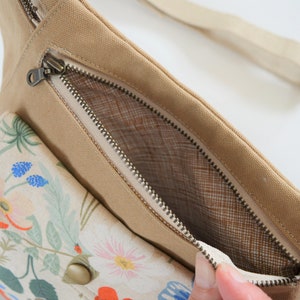 Pu&ograve; includere: Una marsupio in tela beige con una tasca a stampa floreale e uno scomparto principale con cerniera. La cerniera &egrave; aperta, rivelando una fodera in rete marrone.