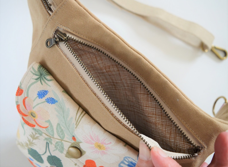 Pu&ograve; includere: Una marsupio in tela beige con una tasca a stampa floreale e uno scomparto principale con cerniera. La cerniera &egrave; aperta, rivelando una fodera in rete marrone.