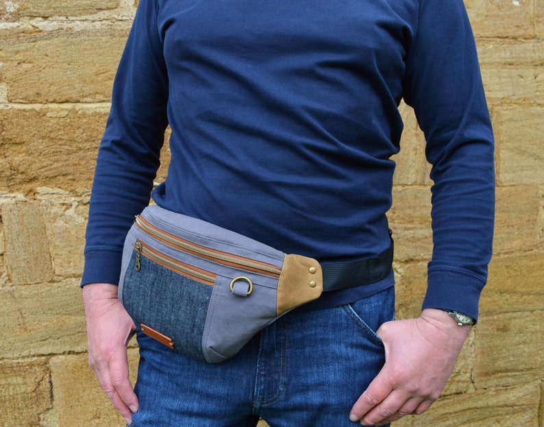 May include: A gray and blue canvas fanny pack with a brown leather flap and a black adjustable strap. The fanny pack has two zippered pockets and a small pocket on the front.