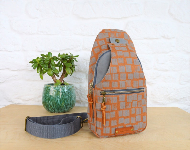 May include: A gray and orange checkered sling bag with a zippered pocket and a leather strap. The bag has a gray fabric lining and a leather patch with the brand name.