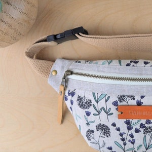 May include: A white canvas fanny pack with a floral print and a brown leather zipper pull. The fanny pack has a beige adjustable strap with a black plastic buckle. The fanny pack has a brown leather tag that says "HANDMADE".
