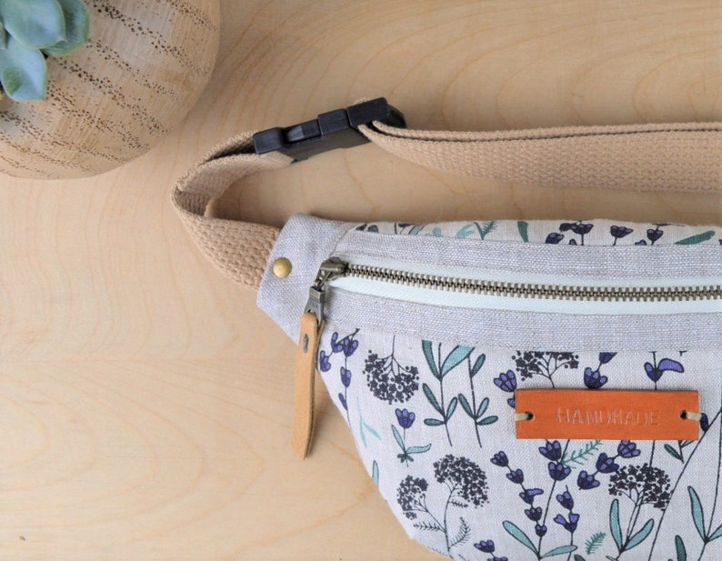 May include: A white canvas fanny pack with a floral print and a brown leather zipper pull. The fanny pack has a beige adjustable strap with a black plastic buckle. The fanny pack has a brown leather tag that says "HANDMADE".