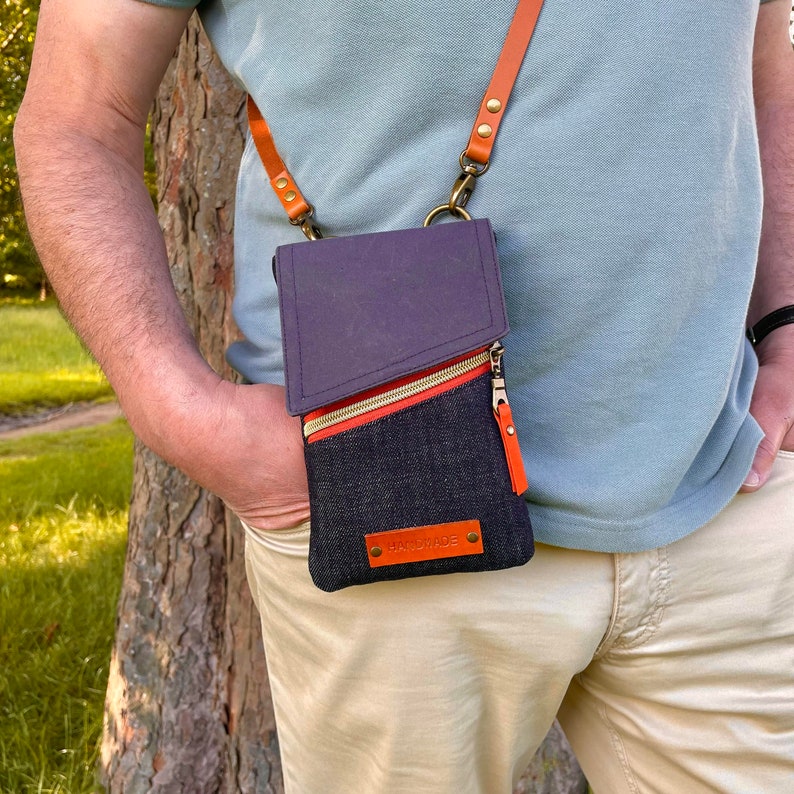 Puede incluir: Un bolso cruzado de mezclilla con solapa morada y cremallera naranja. El bolso tiene una correa de cuero marr&oacute;n y una peque&ntilde;a leng&uuml;eta de cuero naranja. La palabra "HANDMADE" est&aacute; en una peque&ntilde;a etiqueta de cuero naranja.