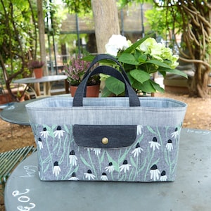 May include: A rectangular tote bag with a gray denim body and black handles. The lower portion features a floral pattern with white flowers and green stems on a gray background. A small pocket with a button closure is on the front.