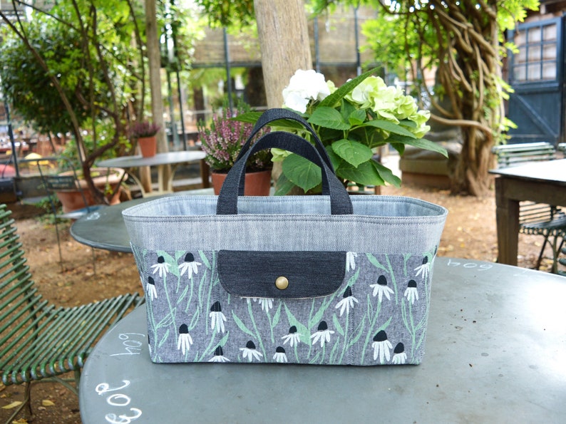 May include: A rectangular tote bag with a gray denim body and black handles. The lower portion features a floral pattern with white flowers and green stems on a gray background. A small pocket with a button closure is on the front.