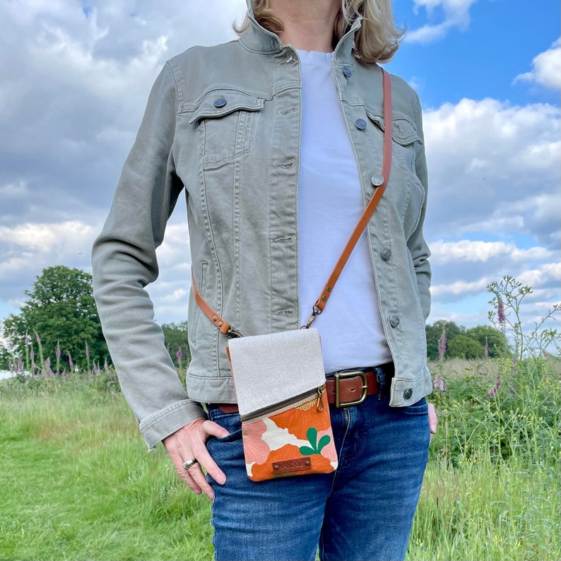 May include: A crossbody phone bag with a tan canvas top and a patterned orange and pink bottom. The bag has a brown leather strap and a gold zipper. The bag is worn over a light green jacket and blue jeans.