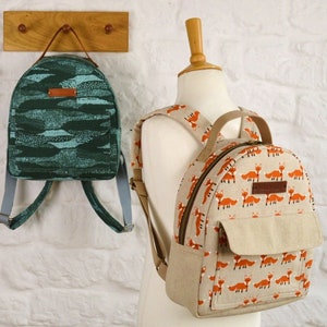 May include: Two backpacks, one green with a wavy pattern and one beige with a repeating fox print. Both backpacks have adjustable straps.