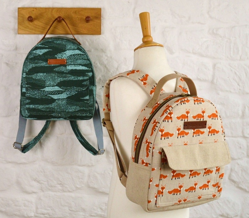 May include: Two backpacks, one green with a wavy pattern and one beige with a repeating fox print. Both backpacks have adjustable straps.