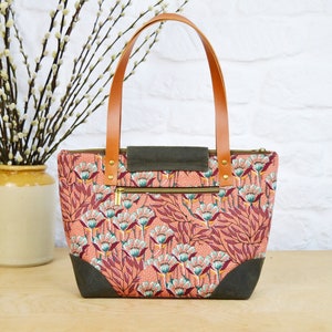 May include: A pink and green floral tote bag with brown leather handles and a zippered pocket. The bag has a dark green canvas lining.