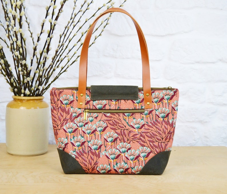 May include: A pink and green floral tote bag with brown leather handles and a zippered pocket. The bag has a dark green canvas lining.
