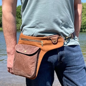Pu&ograve; includere: Una marsupio in pelle marrone con una tasca con cerniera e una chiusura a fibbia. Il marsupio &egrave; indossato in vita da una persona che indossa una maglietta blu chiaro e jeans blu.