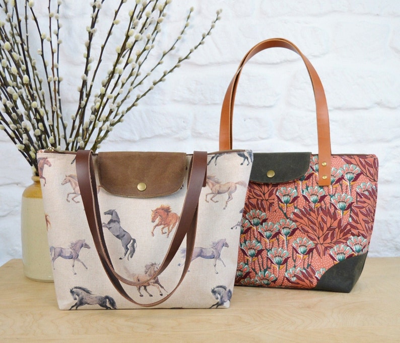 May include: Two tote bags with brown leather handles. One bag is white with a brown suede flap and a horse print. The other bag is red with a floral print and a green suede flap.