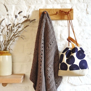 May include: A wooden wall-mounted coat rack with two hooks and a brown crocheted throw draped over one hook. A white tote bag with blue polka dots hangs from the other hook.