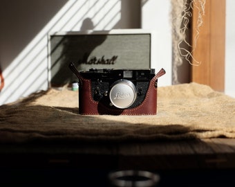 May include: A black camera with a brown leather case and a silver lens cap. The camera is on a textured surface, with a Marshall speaker in the background. The lens cap has the word "Leica" on it.