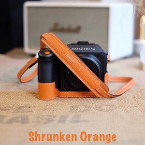 May include: A black camera with a leather case and strap. The case and strap are a bright orange color. The camera has the text "HASSELBLAD" and "X1D" on it. The text "Shrunken Orange" is at the bottom of the image.