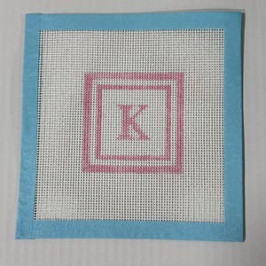 May include: A square needlepoint canvas with a light blue border. The center features a white mesh with a pink square and the letter "K" in the center. The canvas is likely used for embroidery or cross-stitch projects.