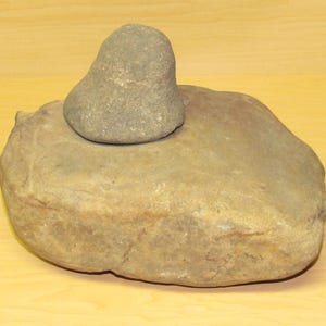 May include: A stone mortar and pestle set. The mortar is a large, rounded stone, and the pestle is a smaller, rounded stone. Both are a light gray color and appear to be made of natural stone. The set is likely used for grinding or crushing ingredients.