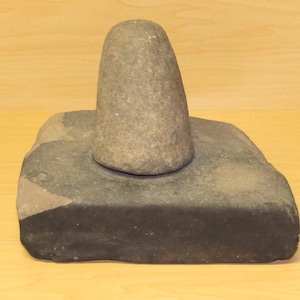 May include: A gray stone mortar and pestle set. The mortar is a rectangular block, and the pestle is a rounded cone shape. The set is made of natural stone and is likely used for grinding ingredients.