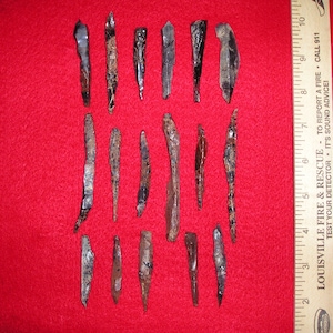 May include: A collection of 16 arrowheads, some with a brown and black mottled pattern, displayed on a red felt surface next to a wooden ruler.