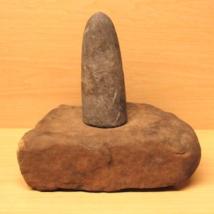 May include: A stone mortar and pestle set. The mortar is a rectangular, reddish-brown stone. The pestle is a gray, elongated stone, standing upright in the center of the mortar. The background is a light brown wooden surface.