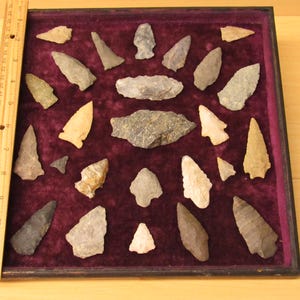 May include: A collection of 25 arrowheads displayed in a velvet-lined wooden box. The arrowheads are various shapes, sizes, and colors, including brown, gray, and white.