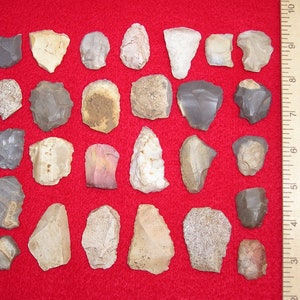 May include: A collection of 25 grey, brown, and white flint arrowheads and spear points on a red felt background. A wooden ruler is in the upper right corner of the image.