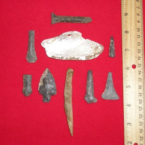 May include: A collection of eight arrowheads and a shell on a red background. The arrowheads are made of various materials, including stone and metal. The shell is white and has a hole in the center. A ruler is visible in the background.