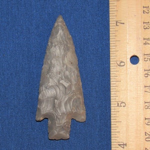 May include: A gray stone arrowhead with a pointed tip and a slightly curved base. The arrowhead is approximately 4 inches long and is shown next to a ruler for scale.