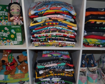 May include: A white shelf unit with three shelves. The top shelf has a stack of colorful t-shirts, the middle shelf has a stack of colorful t-shirts, the bottom shelf has a stack of colorful t-shirts, a pair of brown plush Scooby Doo toys on a green toy car, and a pair of white sneakers with cactus designs. The shelf unit also has two boxes of Perrier water with a floral design.