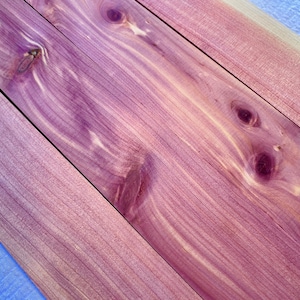 May include: Close-up of four pieces of cedar wood with a reddish-purple hue and a natural wood grain pattern. The wood has a smooth surface and is arranged in a diagonal pattern.