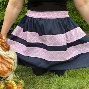 May include: A navy blue and pink apron with a white geometric pattern. The apron has a wide waistband and a gathered skirt.