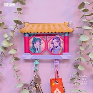 Puede incluir: Un llavero de pared decorativo con un diseño de pagoda, con dos retratos octogonales. El llavero tiene un techo amarillo, columnas rosas y una base lavanda. El texto "NANDEMO CREATIVE" es visible. El llavero tiene dos llaveros.