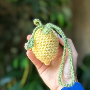 Puede incluir: Un llavero de limón tejido a crochet con un tallo y hojas verdes. El llavero es amarillo y verde y se sostiene en una mano.